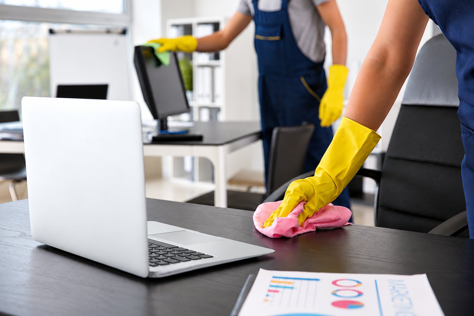 Deux agents d’entretien en salopette bleue et gants jaunes nettoient un bureau noir, un ordinateur portable et un écran dans un bureau lumineux.