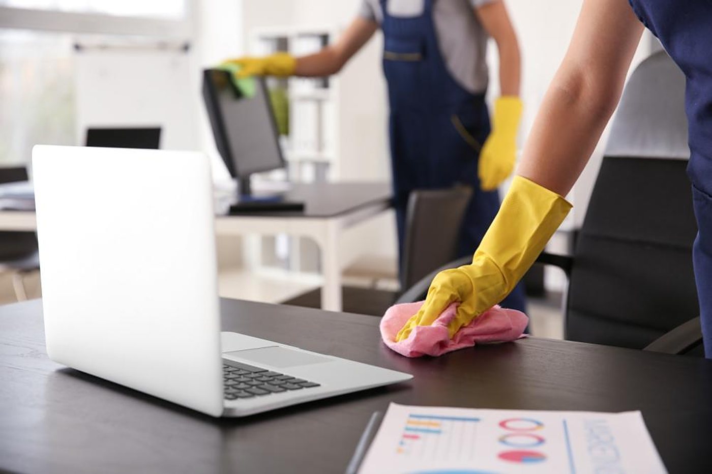 Deux agents de nettoyage en salopette bleue et gants jaunes essuient un bureau et un écran d’ordinateur dans un bureau, avec un chiffon rose au premier plan.